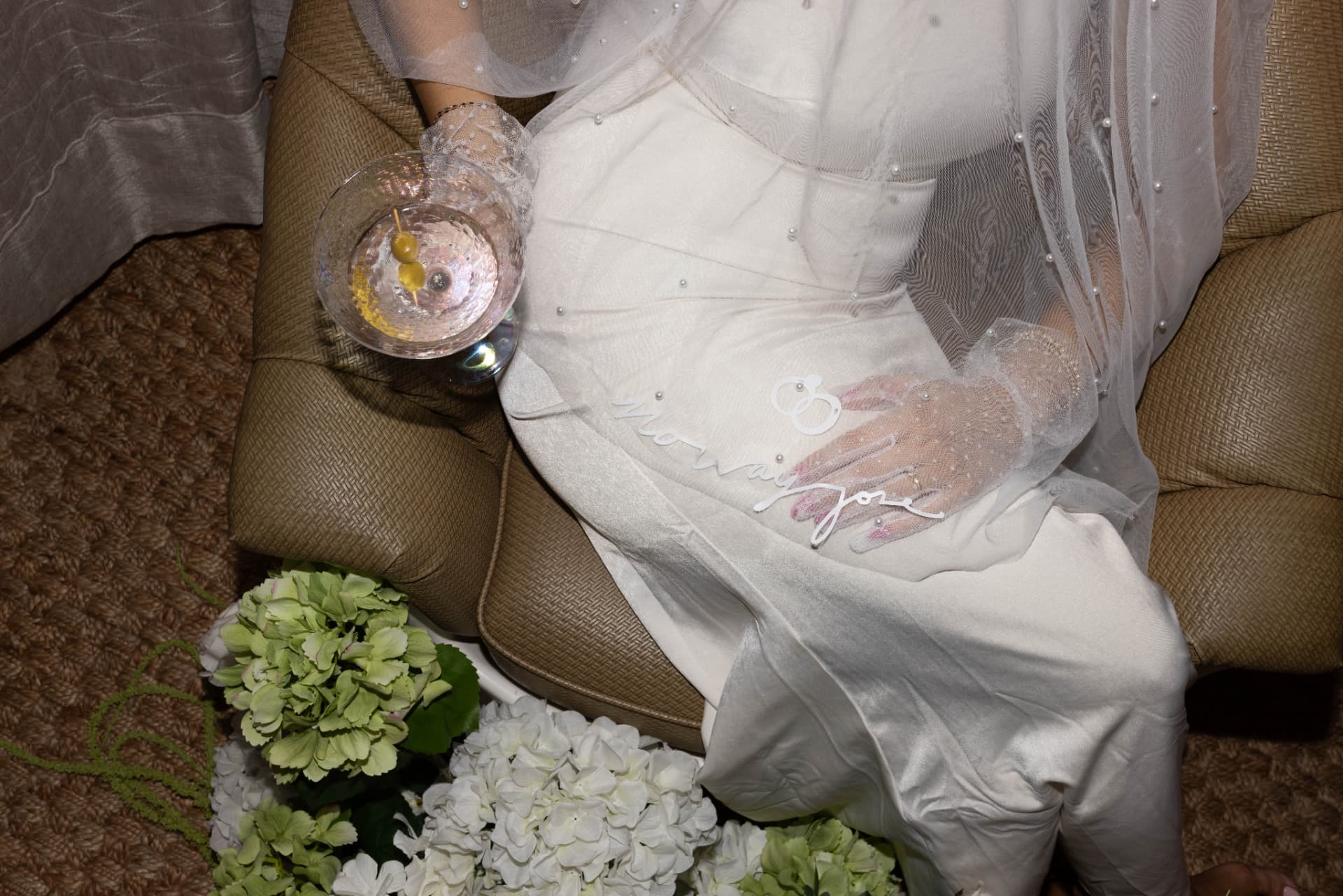 Bride seated with a martini, her no way jose slip dress and veil draped over a woven chair beside hydrangeas